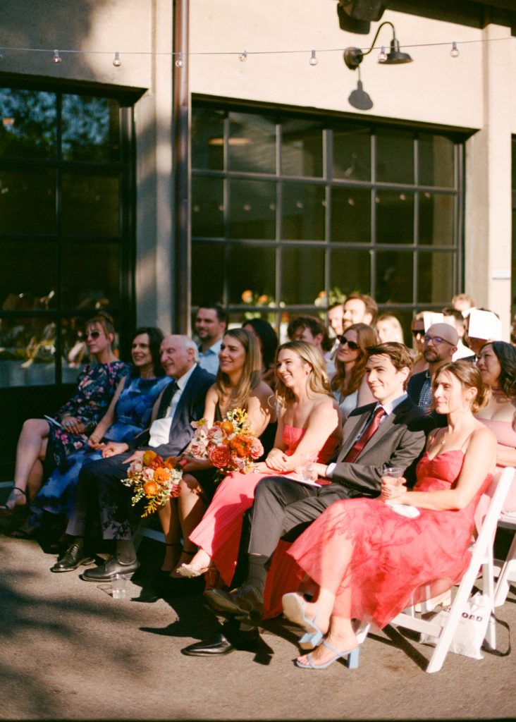 Rows of wedding guests are seated for a ceremony on a patio adjacent to a warehouse-style venue with large glass windows.