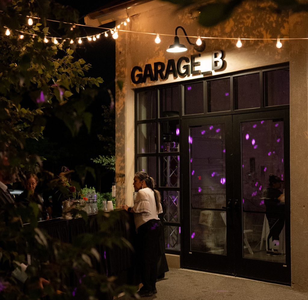 A woman stands at a bar setup underneath string lights on a patio beside Garage B's exterior.
