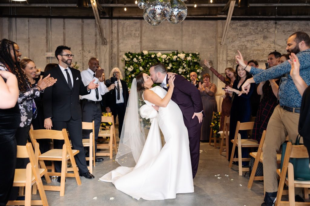 A bride and groom kiss in the middle of the aisle, flanked by rows of cheering loved ones on either side, after their wedding ceremony in a warehouse-style venue.