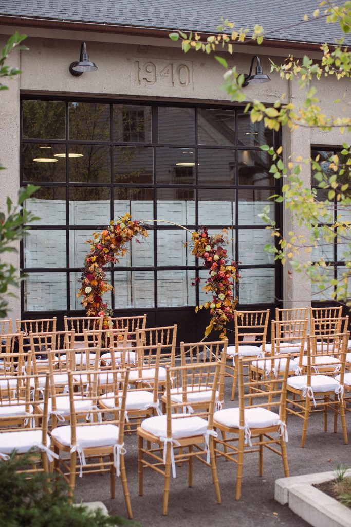 Rows of wooden Chiavari chairs arranged in front of a circular arch featuring autumnal florals before a large glass window at the exterior of a warehouse-style wedding venue.