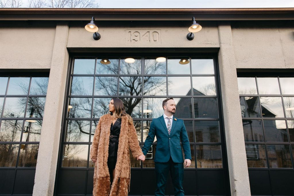 A woman and man stand hand-in-hand, looking away from each other, in front of a large glass window on the side of a building established in 1940