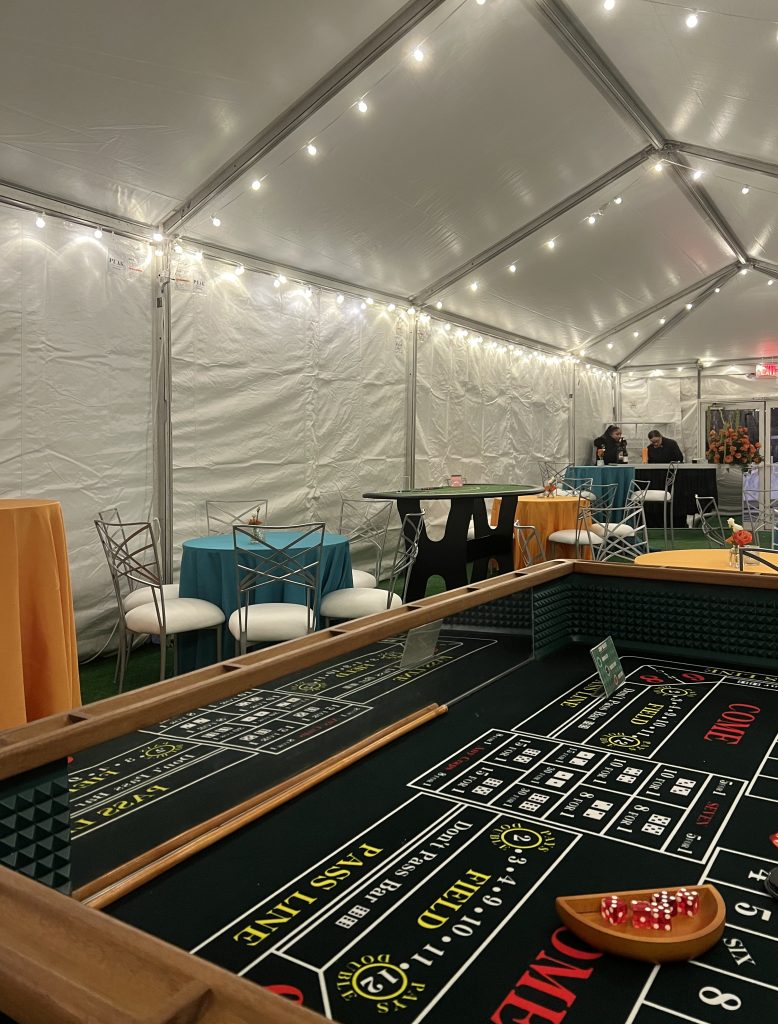 A tented space outfitted with casino games, a bar, tables, chairs, and string lights at the ceiling.