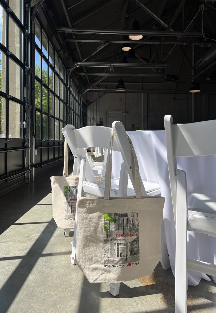 A tote bag, featuring a sketched graphic of a New Balance storefront, hanging on a folding chair at a banquet table inside a warehouse-style venue with steel beams at the ceiling and large glass windows.