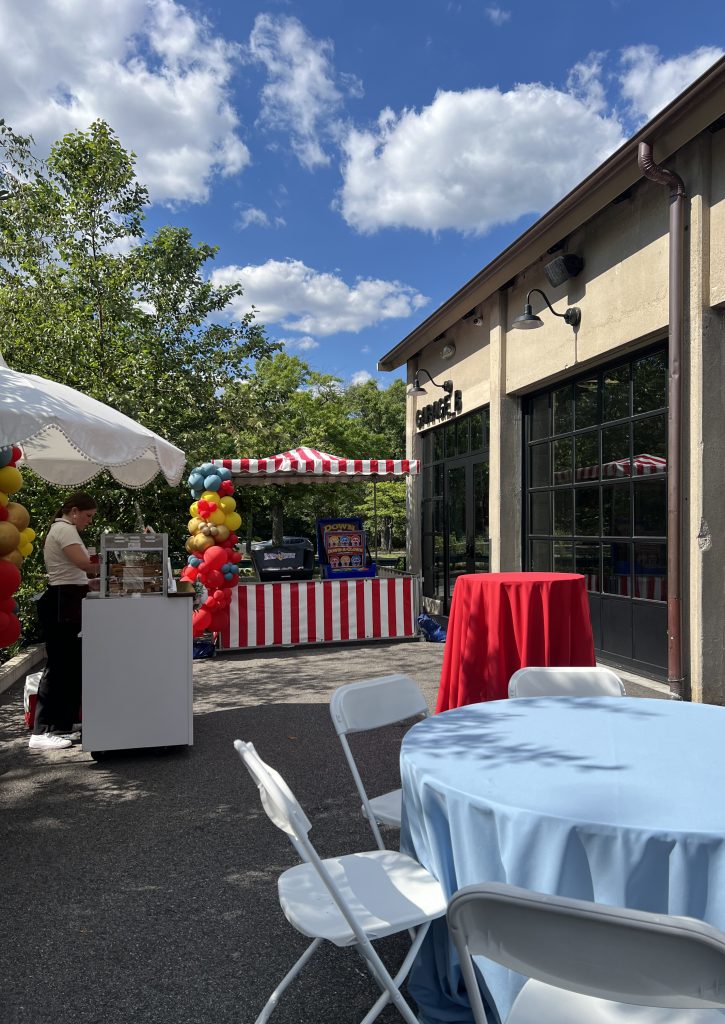 An arrangement of tables, a snack station, and a carnival game on the patio adjacent to Garage B's exterior.