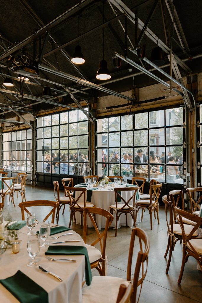 A view from inside a modern-industrial wedding venue, outfitted with banquet tables and wooden X-back chairs, through a bank of floor-to-ceiling glass windows to a crowd of wedding guests mingling outdoors.
