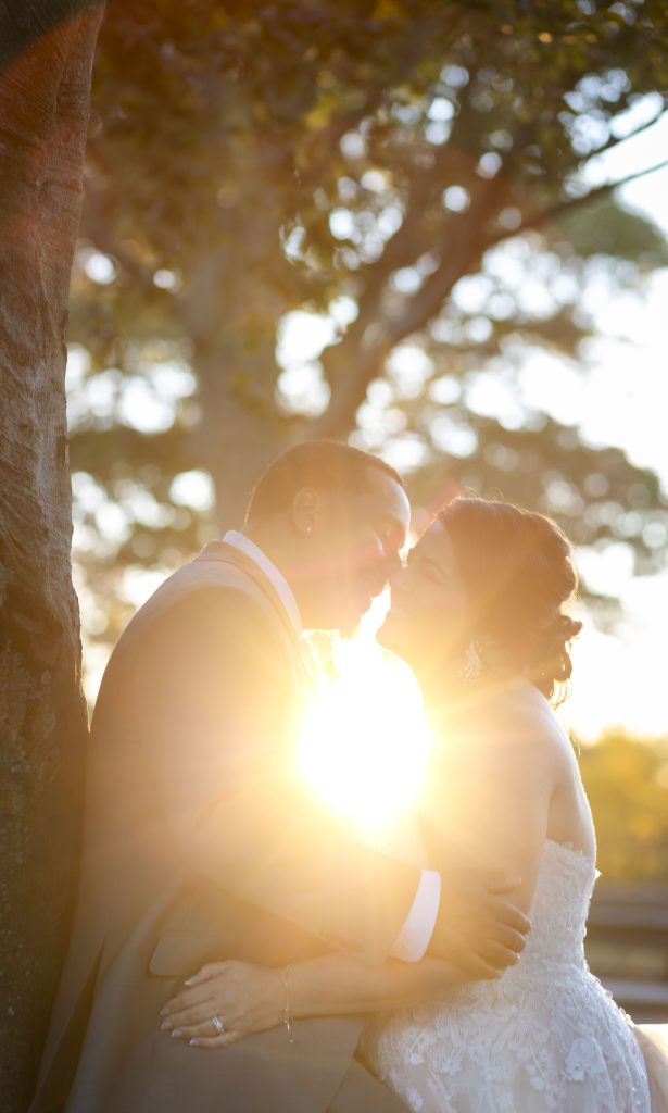A bride and groom smiling and nearly kissing by a tree as the sunset shines from behind them.