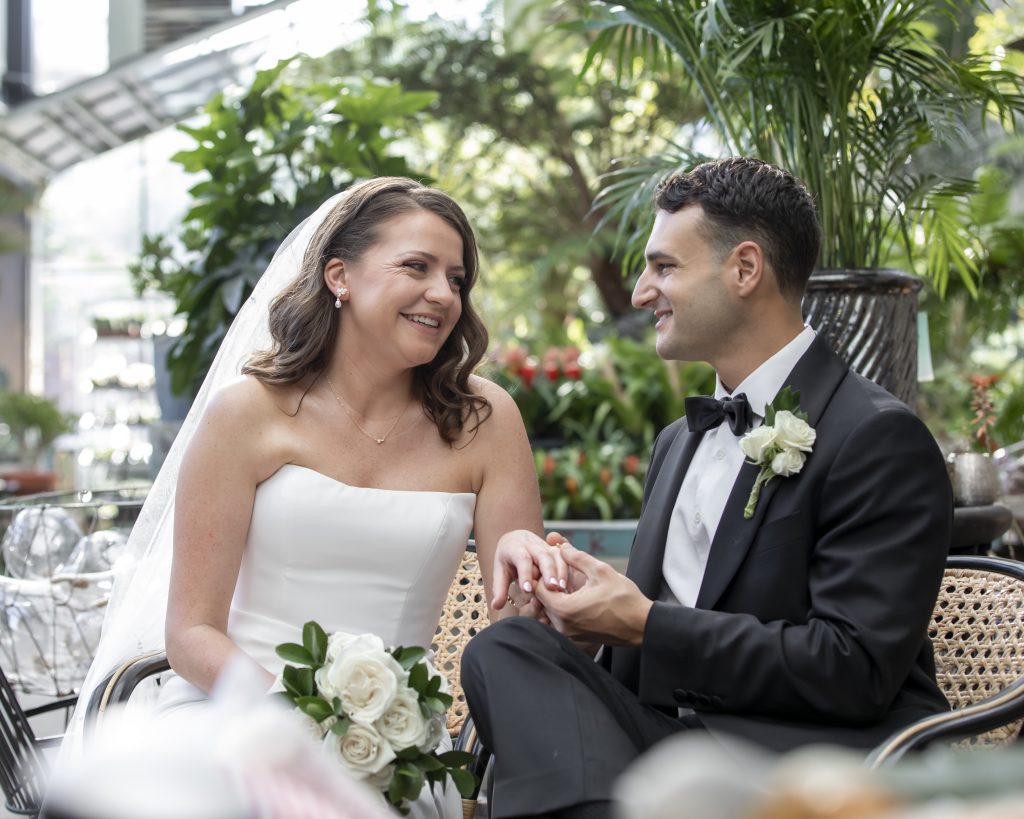A bride and groom, sitting on a bench in a garden center, look at each other while smiling.