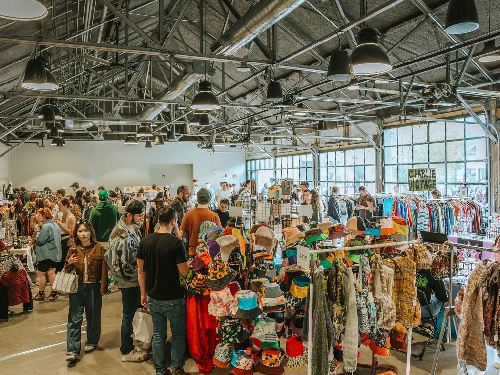 A vintage clothing market inside a modern-industrial event space featuring high ceilings, steel trusses, pendant lighting, and a bank of large glass windows.