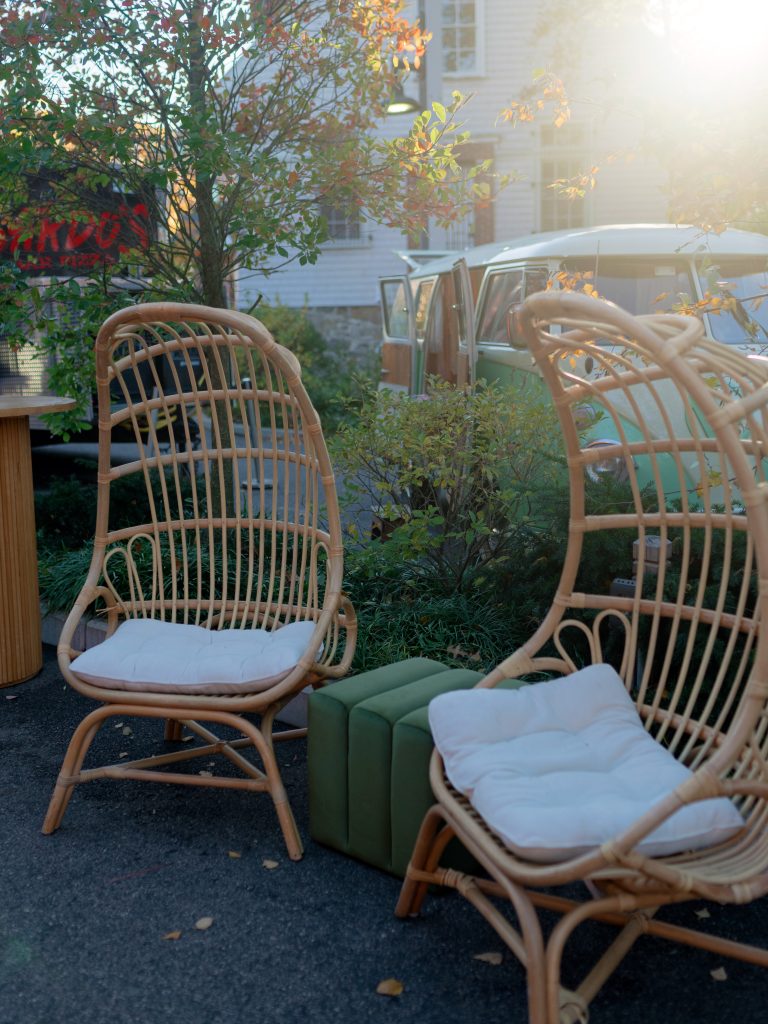 A lounge area, consisting of two wicker high-back chairs and a cushion, on a patio with a food truck and a vintage Volkswagen van in the background.
