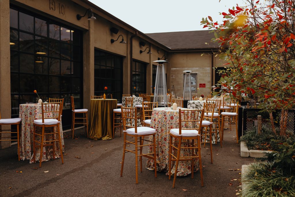 A patio beside a modern-industrial venue, featuring a bank of large glass windows, outfitted with high-top tables and chairs as well as stand-up propane heaters.