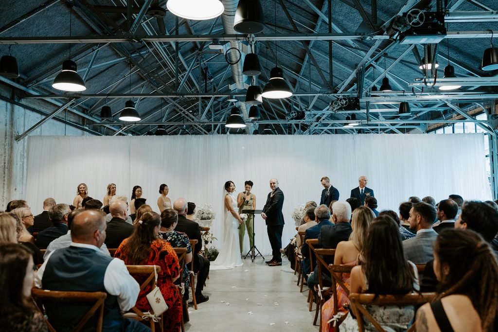 A wedding ceremony takes place in front of a wall-to-wall drape partition underneath a ceiling feature steel rafters and large pendant lights.