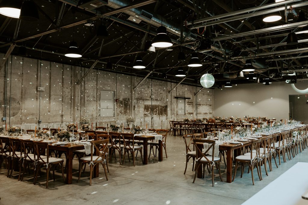 An arrangement of farm tables set up for a wedding reception underneath a disco ball hung from steel rafters.
