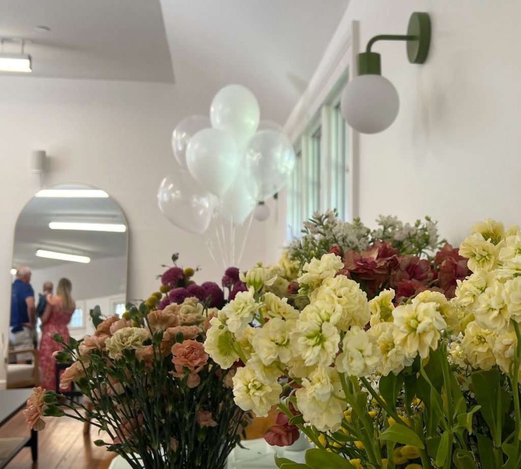 A white room with bouquets of flowers in the foreground and a floor-length mirror in the background that shows the reflection of two people holding a baby.