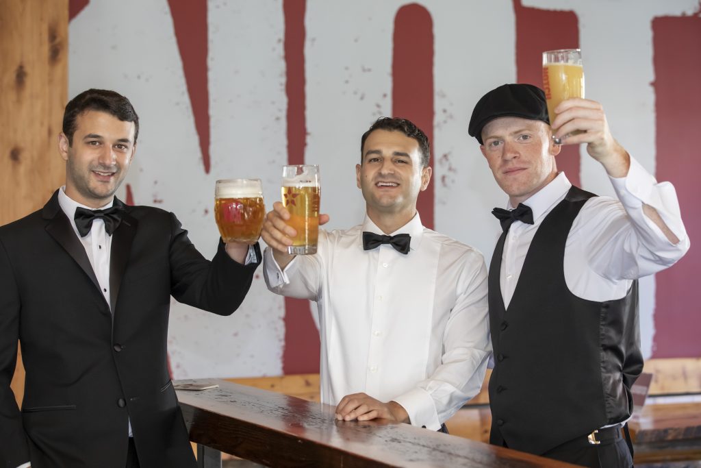 Three groomsmen toast to the camera with pints of beer inside the Brighton location of Notch Brewing in Boston, Massachusetts.