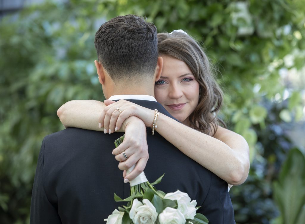 A bride, facing the camera, hugs her groom, turned away from the camera, against a background of greenery.