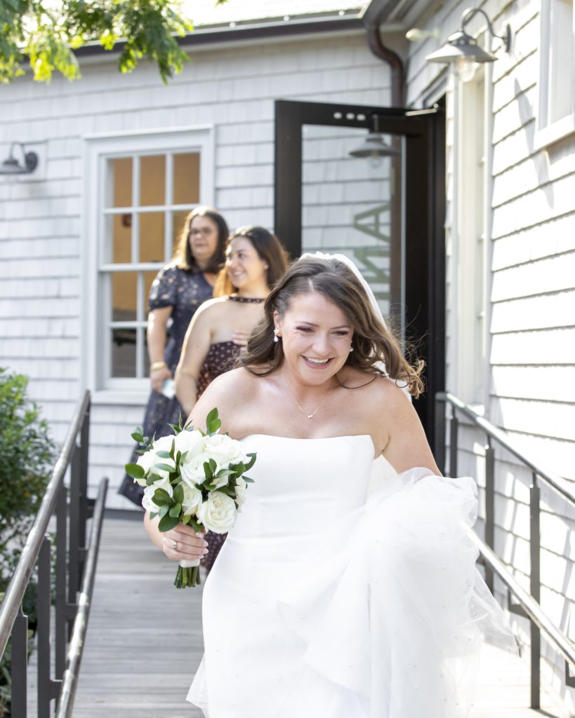 A bride, holding a bouquet and followed by two members of her bridal party, walk down a ramp as they leave a Shingle and Colonial Revival style building.