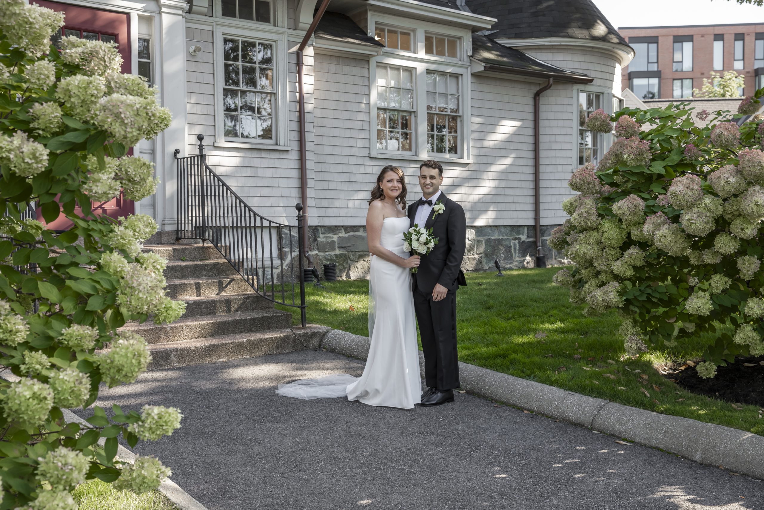 A bride, holding a bouquet, and a groom stand on a pathway flanked by hydrangea bushes in front of a Shingle and Colonial Revival style building.