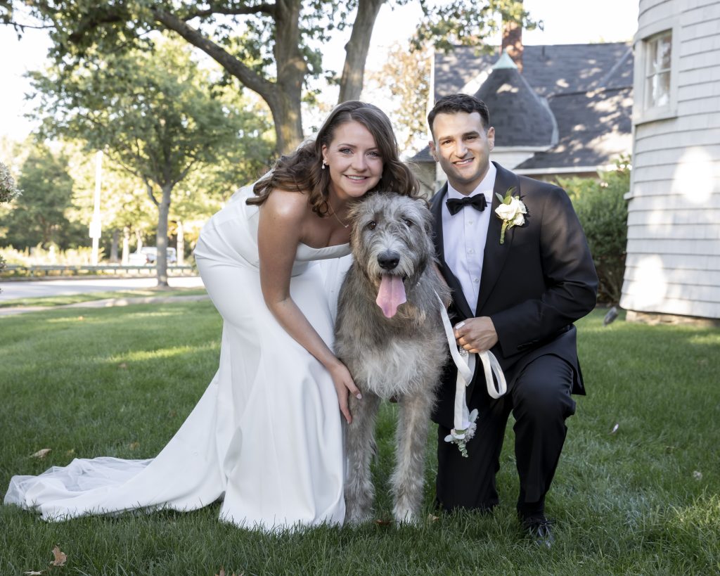 A bride and groom pose, squatting, with an Irish wolfhound in a grassy yard with a Shingle and Colonial Revival building in the background.