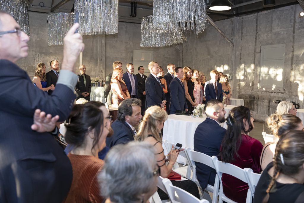 Standing and seated wedding guests fill a modern-industrial venue outfitted with silver streamers at the ceiling.