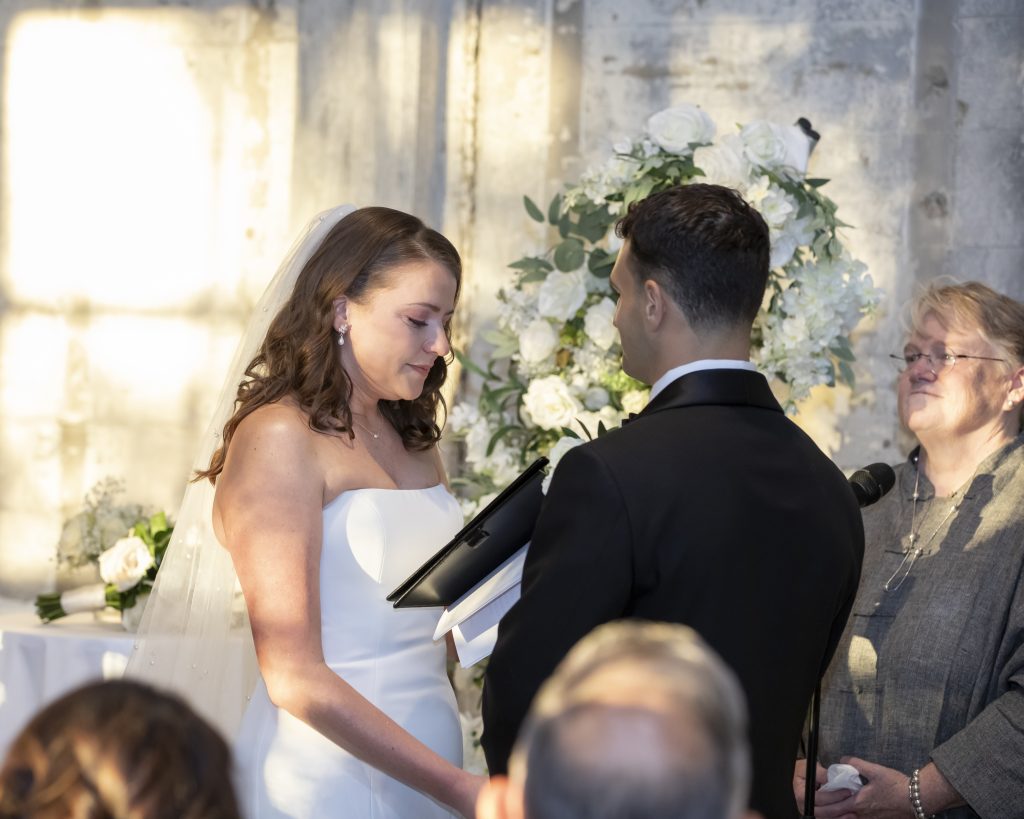 Sunlight washes over a bride reading her vows to her groom and their officiant against a distressed stone wall.