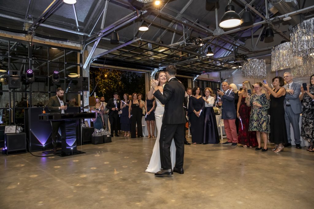 A bride and groom share their first dance surrounded by loved ones in a warehouse-style venue that features an opened bank of glass floor-to-ceiling windows, steel rafters, pendant lighting, and polished concrete flooring.