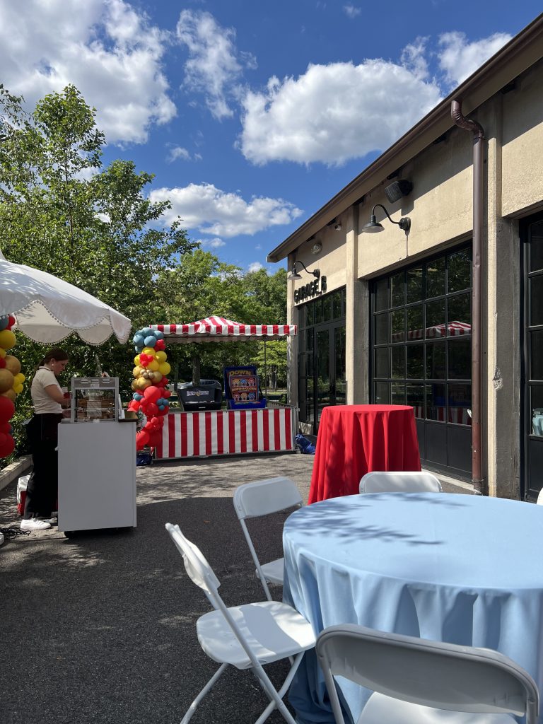 The outdoor patio adjacent to Garage B, outfitted with a low-top and high-top table, a game station, and a dessert cart, all designed in a carnival theme.