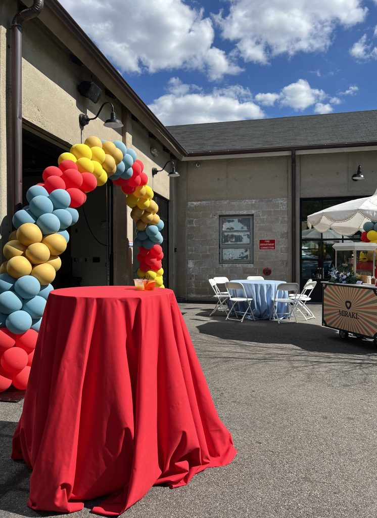 A patio adjacent to a warehouse-style building with a balloon arch at its entrance, outfitted with a low-top and high-top table and a dessert cart designed in a carnival theme.