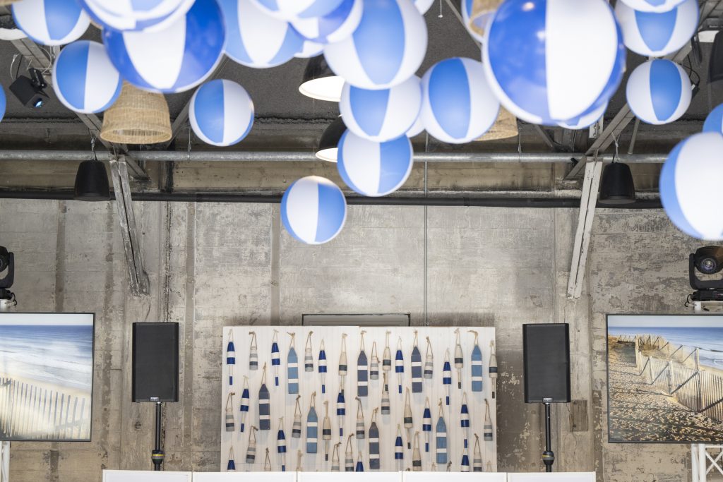 Beach-themed decor in a venue that features steel rafters and distressed stone walls, including beach balls suspended at the ceiling and a DJ booth with a backdrop comprised of buoys and monitors displaying seaside vistas.