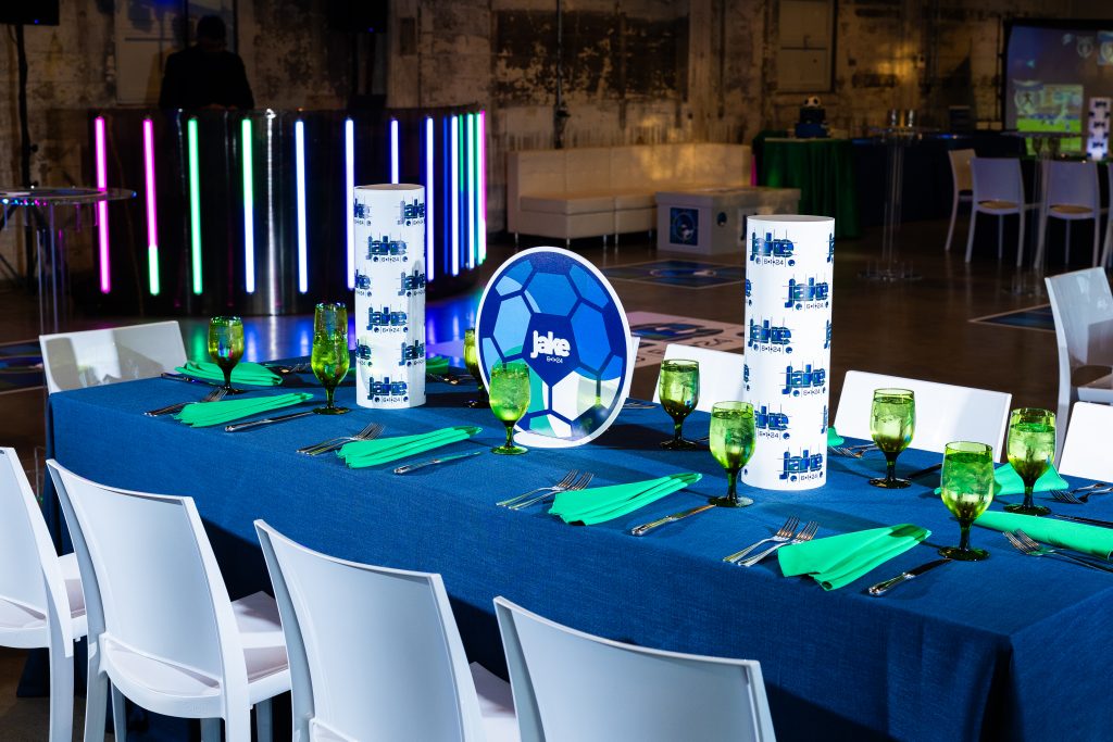 A rectangular banquet table at a soccer-themed bar mitzvah reception with a DJ booth, lounge seating, and soccer simulator game in the background.
