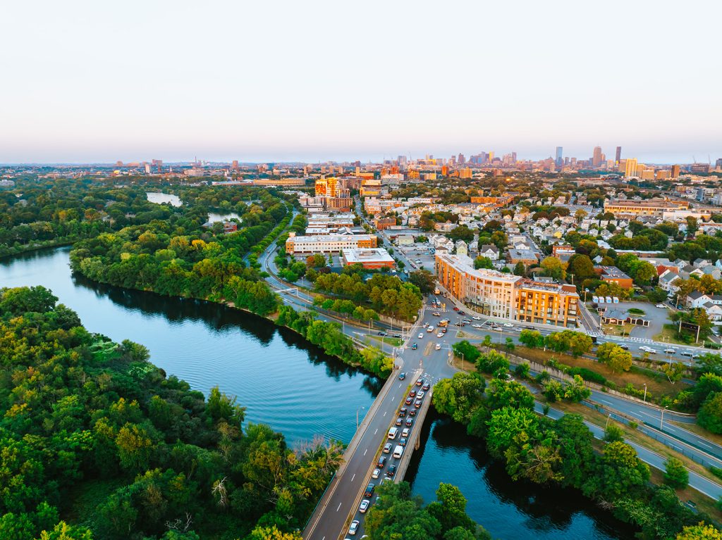 An aerial view at sunset of the northwest corner of the Brighton neighborhood of Boston, featuring a bend of the Charles River and Soldiers Field Road in the foreground and the city skyline in the background.