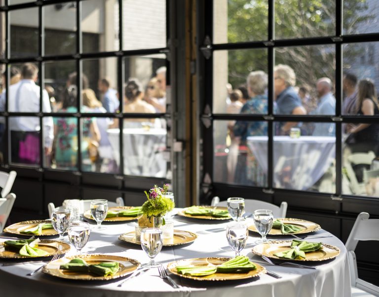 A view through large glass windows to an outdoor patio space populated with people mingling around high-top tables with a round banquet table set with dinnerware in the foreground.