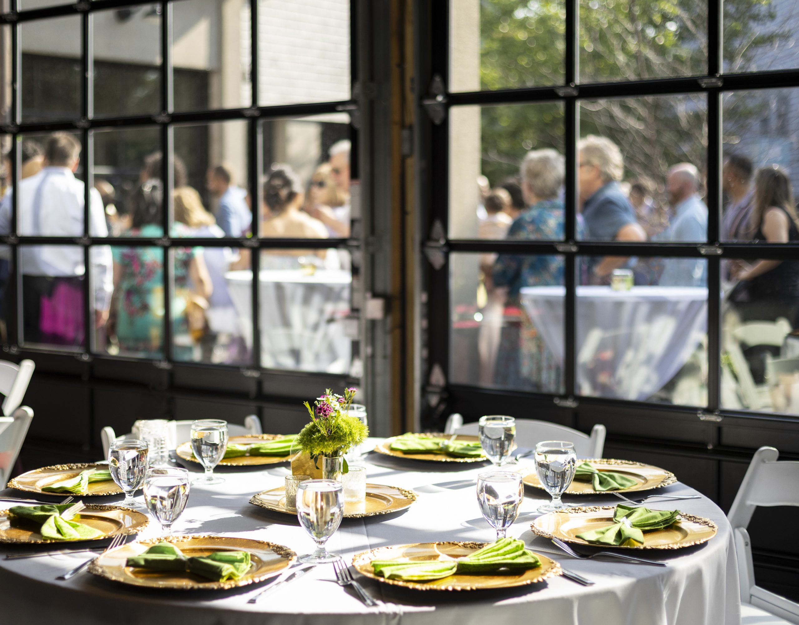 A view through large glass windows to an outdoor patio space populated with people mingling around high-top tables with a round banquet table set with dinnerware in the foreground.