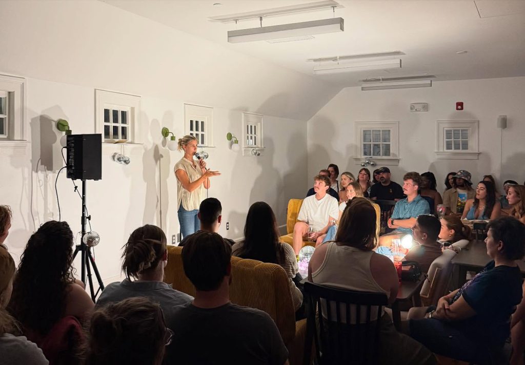 A woman speaks on a microphone in front of an intimately-sized audience sitting on lounge furniture in a micro venue that features cottage-style windows and white walls.