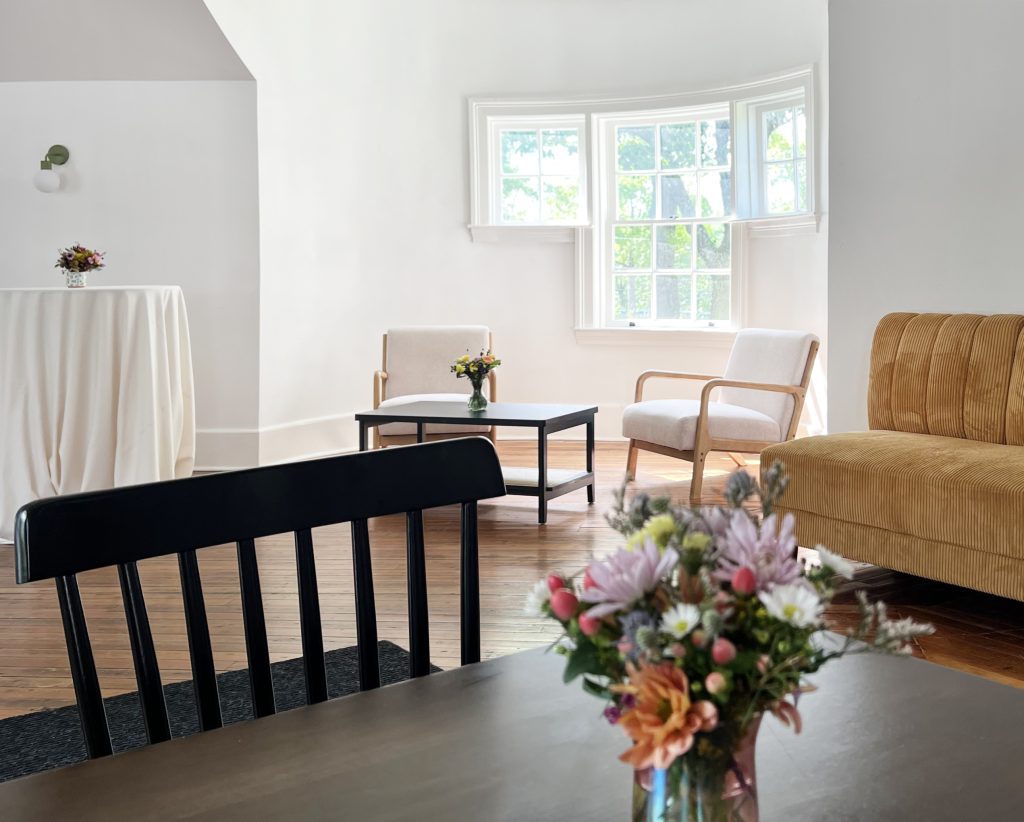 An arrangement of lounge furniture in a room with white walls, cottage-style windows in a rounded turret corner, and hardwood floors, sparsely decorated with small vases of wildflowers.
