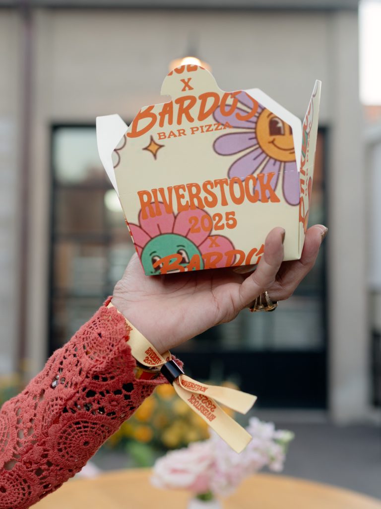 A hand holds a small to-go box of food customized with the branding of the caterer and the name of the event.