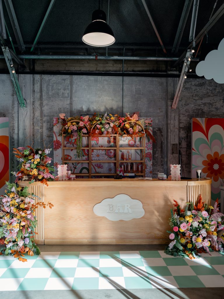 A wooden bar setup adorned with large floral arrangements and fronted by checkered vinyl flooring against a distressed stone wall.