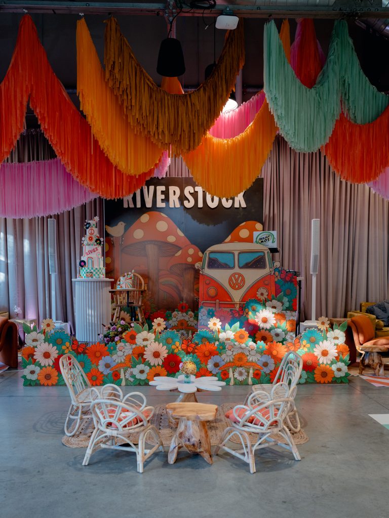 A six-layered cake, a high chair, and a DJ booth styled after a retro Volkswagen van set against a draped wall underneath arches of colorful streamers with an arrangement of children's rattan furniture in the foreground.