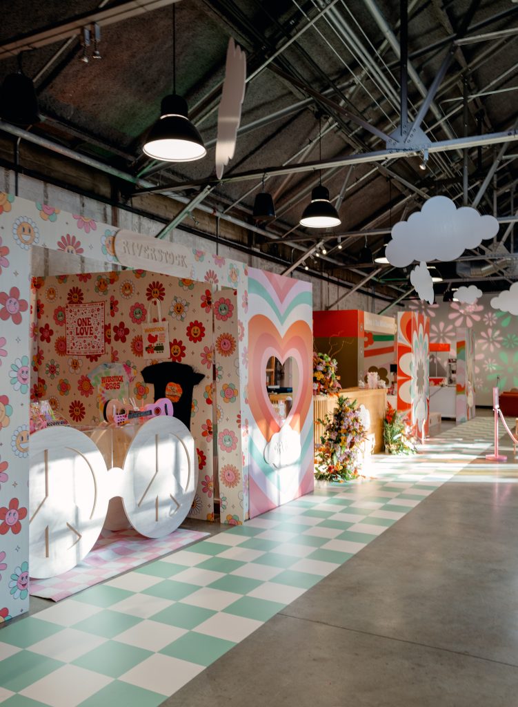 In a warehouse-style venue with steel beams, pendant lighting, and polished concrete flooring, a row of custom-built stalls features a merchandise booth, a bar, and a cotton candy stand.