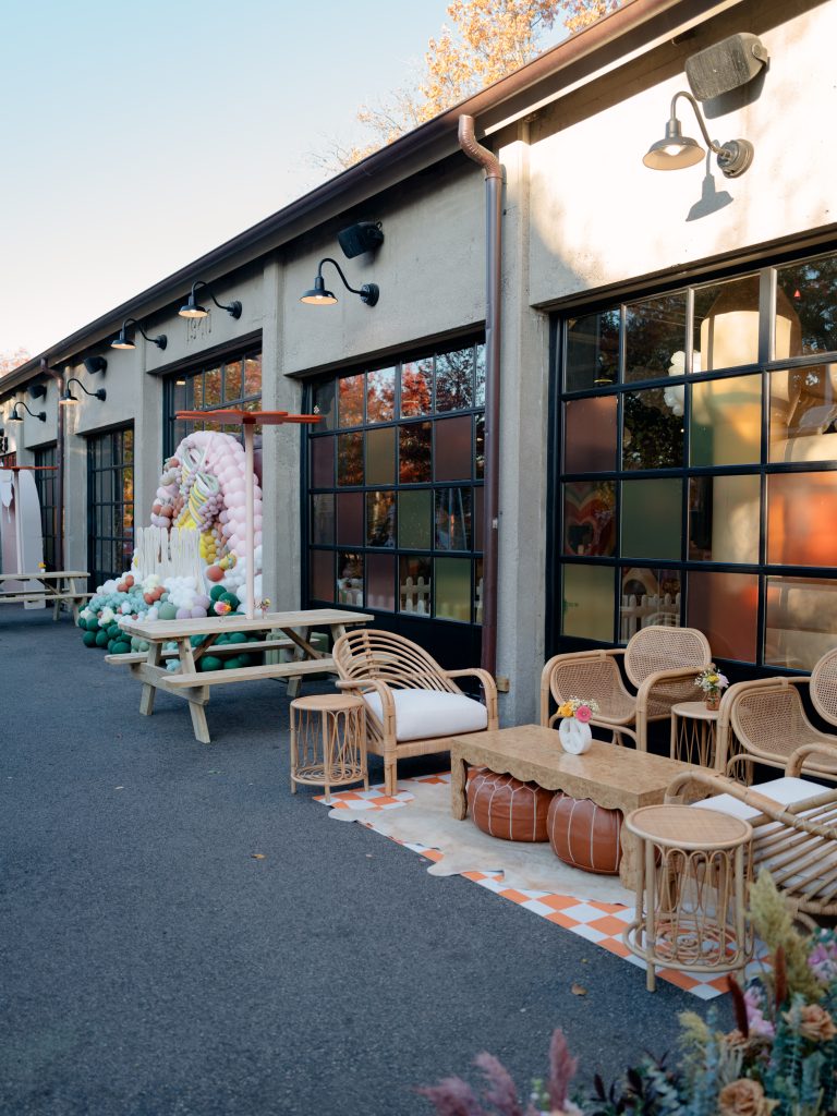 The exterior of Garage B, featuring the bank of large glass windows decorated with colorful film, and the adjacent patio, arranged with rattan lounge furniture, picnic tables, and a balloon arch.