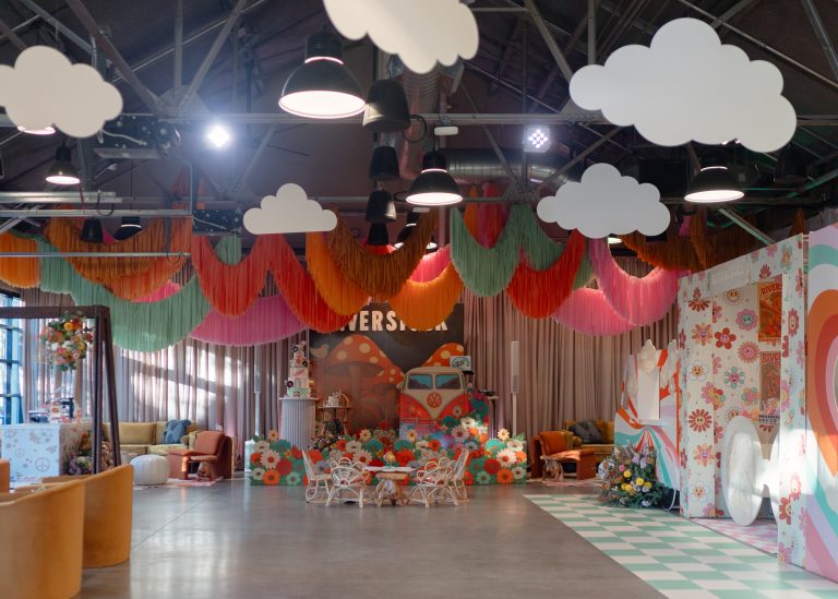 A modern-industrial wedding venue, featuring steel rafters and pendant lighting at the ceiling, decorated for a birthday party with a hippie music festival theme, including streamers, drapery, and lounge furniture.