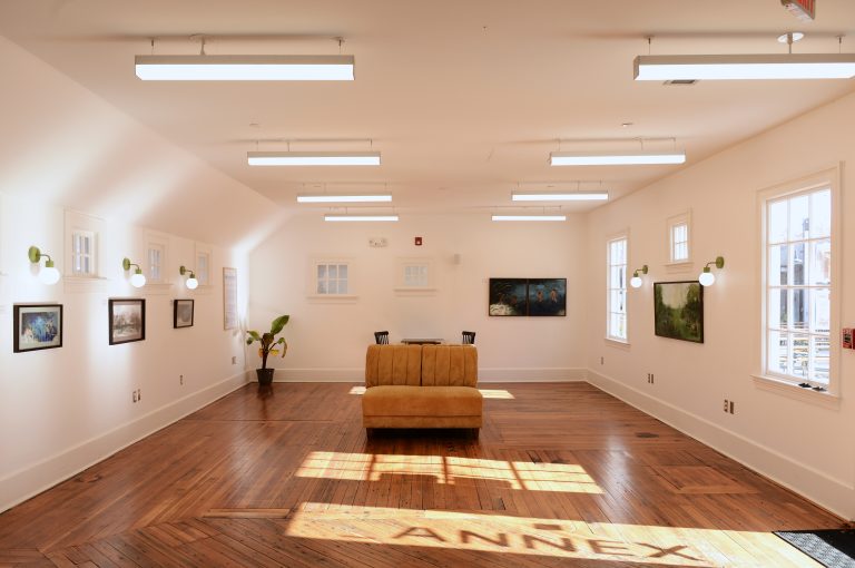 A view of The Annex, outfitted with sparse lounge furniture and paintings hung on the white walls, featuring a shadow of the microvenue's branding on the hardwood flooring.