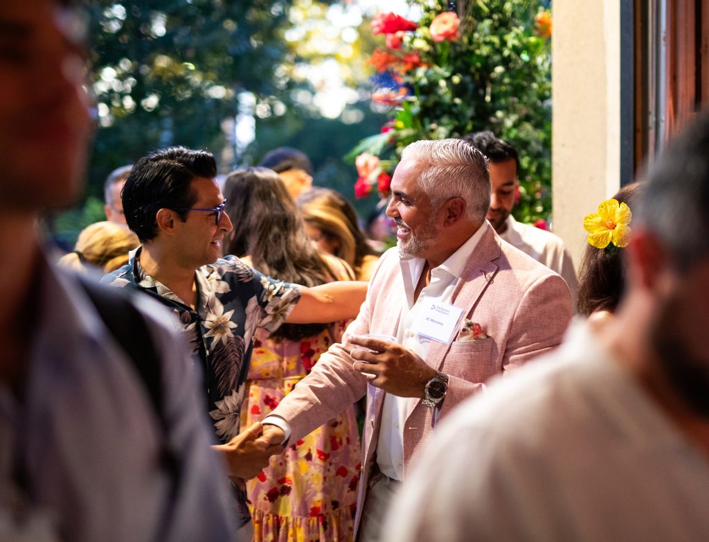 Two people shake hands in the indoor-outdoor threshold of a venue wherein which others are mingling during a cocktail party.