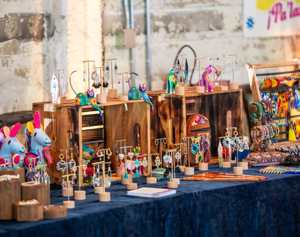 A rectangular table topped with colorful hand-made Mexican jewelry and other accessories, arranged among wooden display boxes, set against a distressed stone wall.