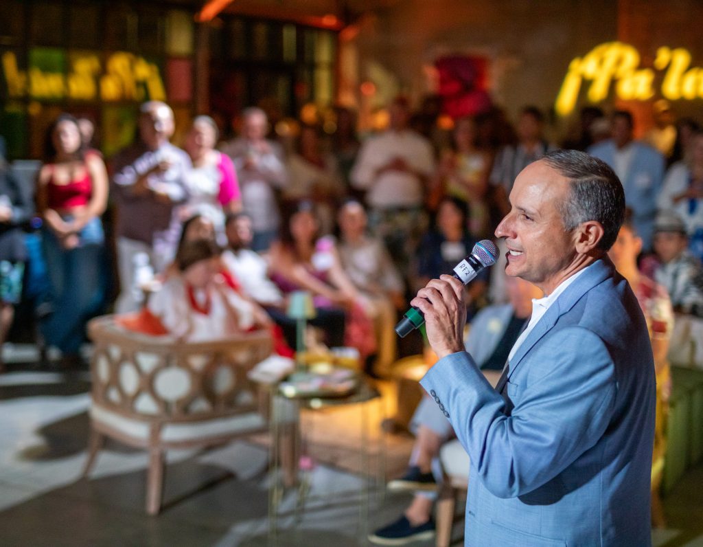 In a warehouse-style venue featuring warm uplighting, a person speaks on a wireless microphone in front of a crowd of sitting and standing people.