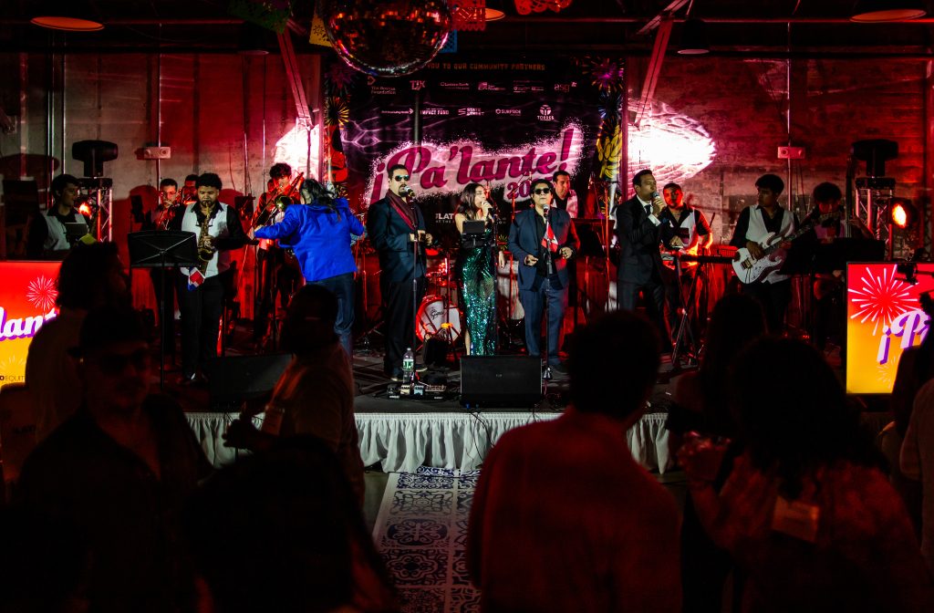 A 15-member house band performs on a stage set against a backdrop printed with the phrase "¡Pa'lante!" in front of a dancing crowd in silhouette.