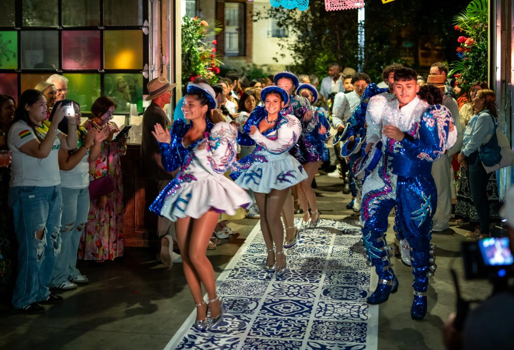 Caporales performers enter a venue upon patterned vinyl flooring from an outdoor patio through an open overhead door to onlooking event guests.