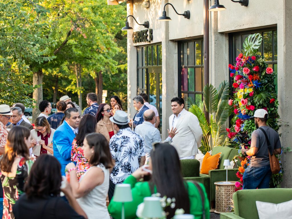 People mingle as part of a cocktail party on an outdoor landscaped patio next to Garage B that is outfitted with lounge areas and tropical floral arrangements.