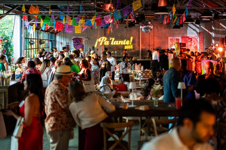 A warehouse-style venue filled with attendees of a cultural gala that features two bar setups against a bank of glass overhead doors, centralized lounge areas, and a stage upon which a house band performs under a disco ball and papel picado hung from steel rafters at the ceiling.