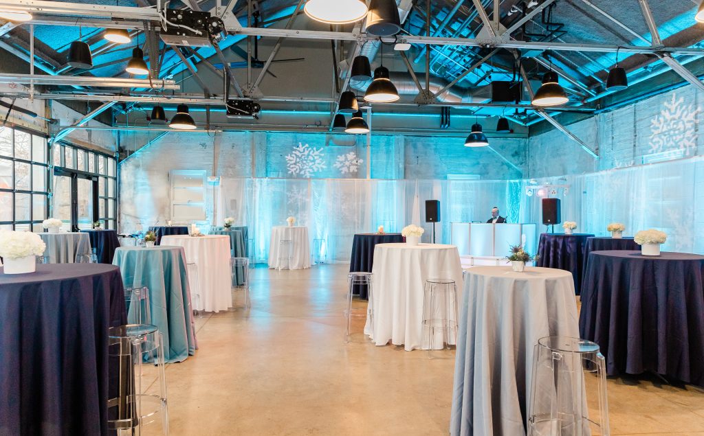 A warehouse-style venue -- featuring steel rafters, distressed stone walls, and polished concrete flooring -- set up for a cocktail party with a pipe-and-drape installation lining the walls, high-top tables dressed with linens and small floral centerpieces, a DJ booth in a far corner, and blue uplighting and snowflake gobo lighting.