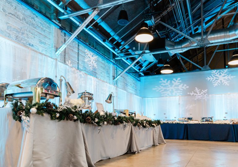 A low angle of a warehouse-style venue -- featuring steel rafters, pendant lighting, distressed stone walls, and concrete flooring -- lined with a pipe-and-drape installation, lit with blue uplighting and snowflake gobo lighting, and outfitted with long banquet tables topped with buffet fare.